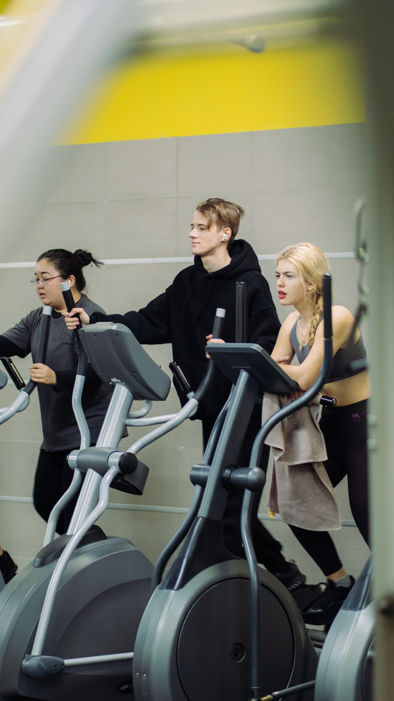 Three people are exercising on elliptical machines in a gym, wearing athletic clothing and using headphones.