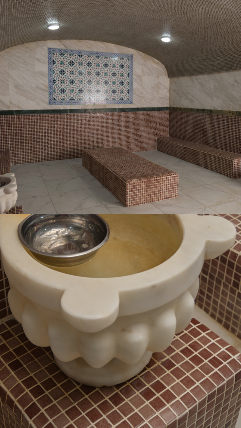 The image shows the interior of a hammam, featuring tiled benches, walls, and a mosaic artwork, along with a marble washbasin containing a metal bowl.