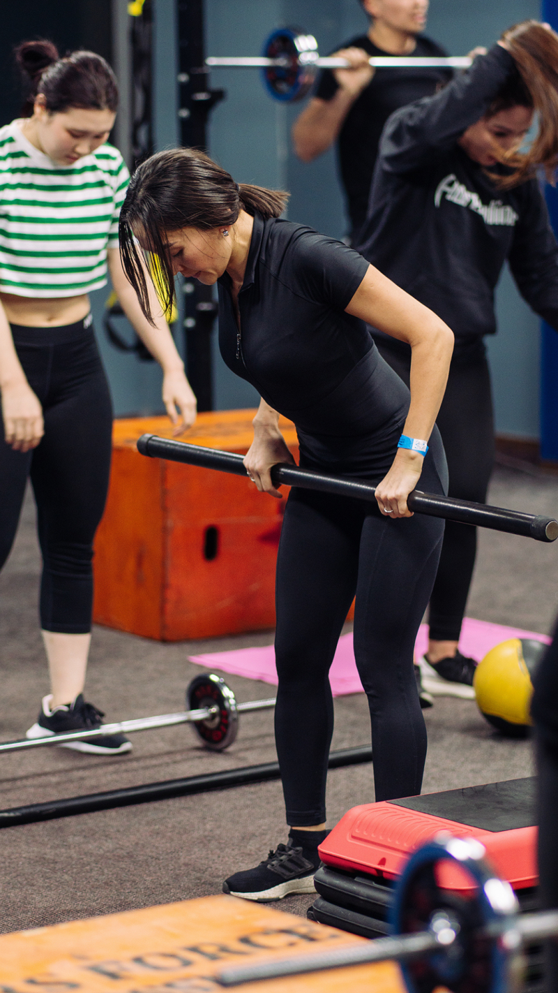 In a gym setting, a woman in black athletic wear is bending over while holding a black bar, with other people and exercise equipment visible in the background.