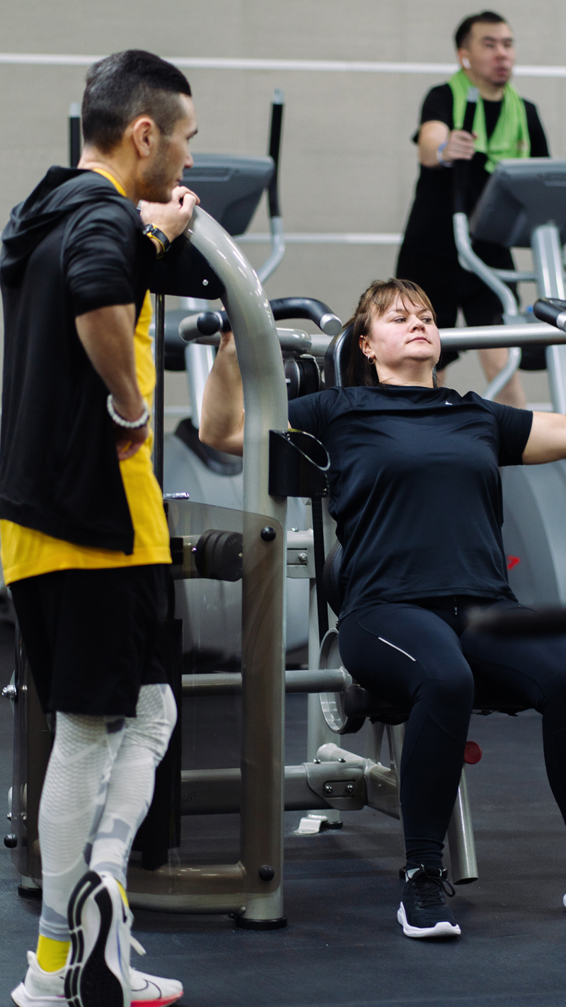 In a gym, a woman is using a machine while a man stands nearby, seemingly assisting or observing; another person is visible in the background using an elliptical machine.