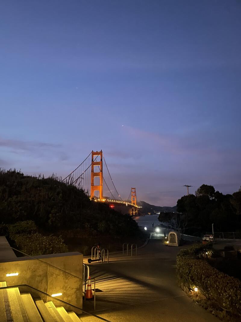 The Golden Gate Bridge is lit up at night, seen from an elevated viewpoint with stairs and a path leading towards it.
