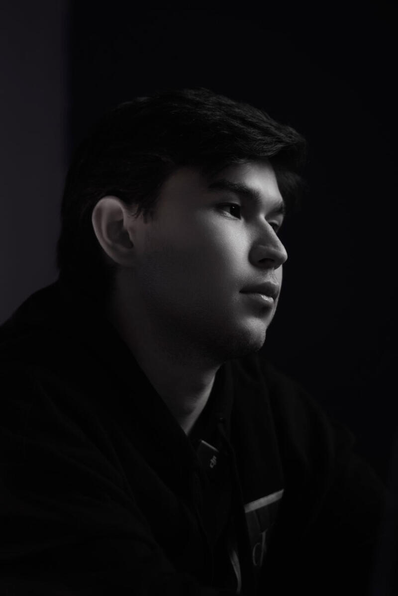 A black-and-white portrait of a young man with dark hair, looking to the right, wearing a dark shirt.