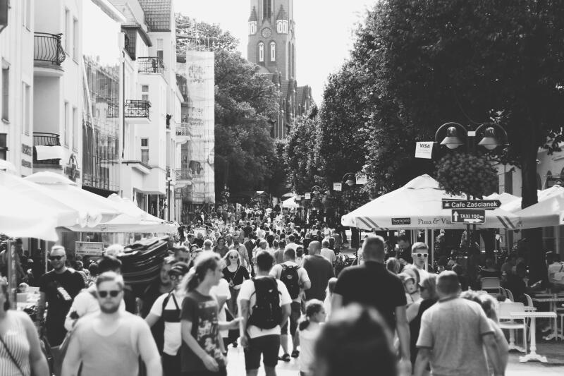 A black and white shot of a busy street with many people walking, lined with buildings and trees, with a church tower visible in the distance. Signage for 'Cafe Zascianek' and 'Taxi 100m' can be seen.