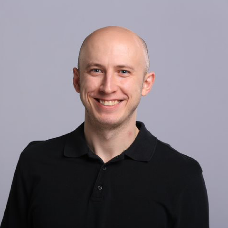 A bald, light-skinned man with blue eyes and a wide smile is wearing a black polo shirt against a light gray background.