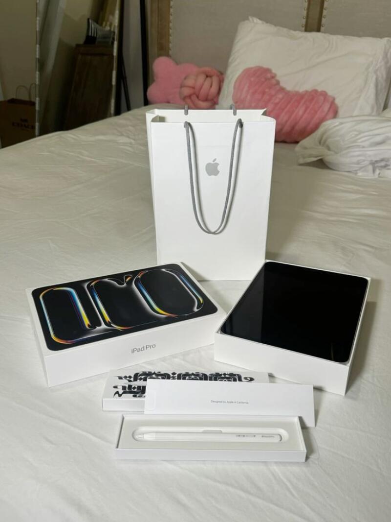 The image shows an arrangement of Apple products on a white bed, including an iPad Pro box, an open iPad box with the screen visible, a white Apple Pencil in its packaging, and an Apple shopping bag.