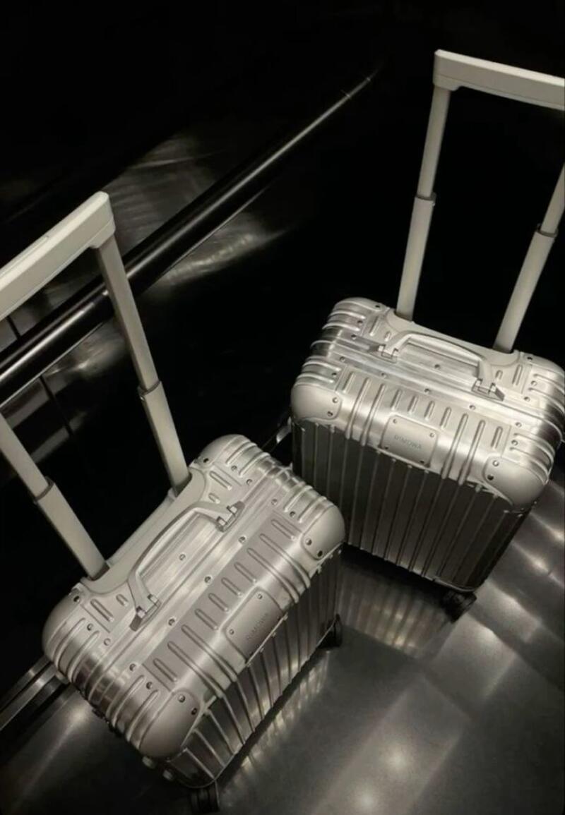 Two silver Rimowa suitcases are positioned on a dark, reflective surface, with their handles extended upwards.