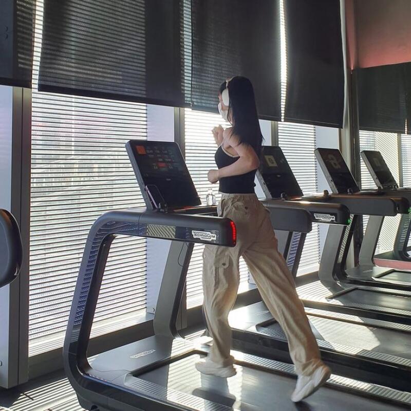 A woman wearing a face mask and headphones is running on a treadmill in a gym, next to a window with closed blinds.