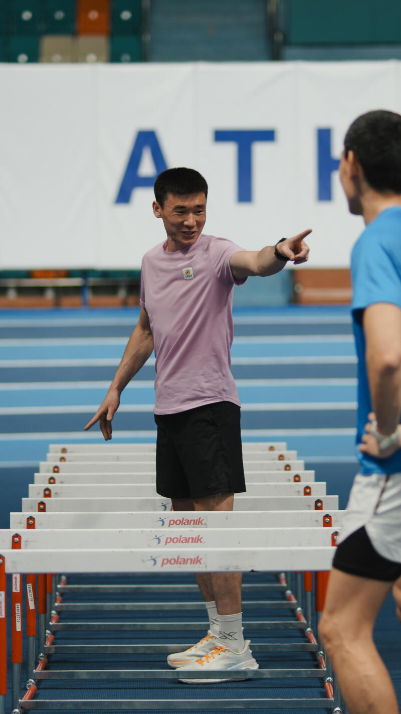 A man in a pink shirt and black shorts stands behind hurdles and points at another man in a blue shirt; the hurdles have the word "polanik" printed on them, and the backdrop features the letters "ATH."