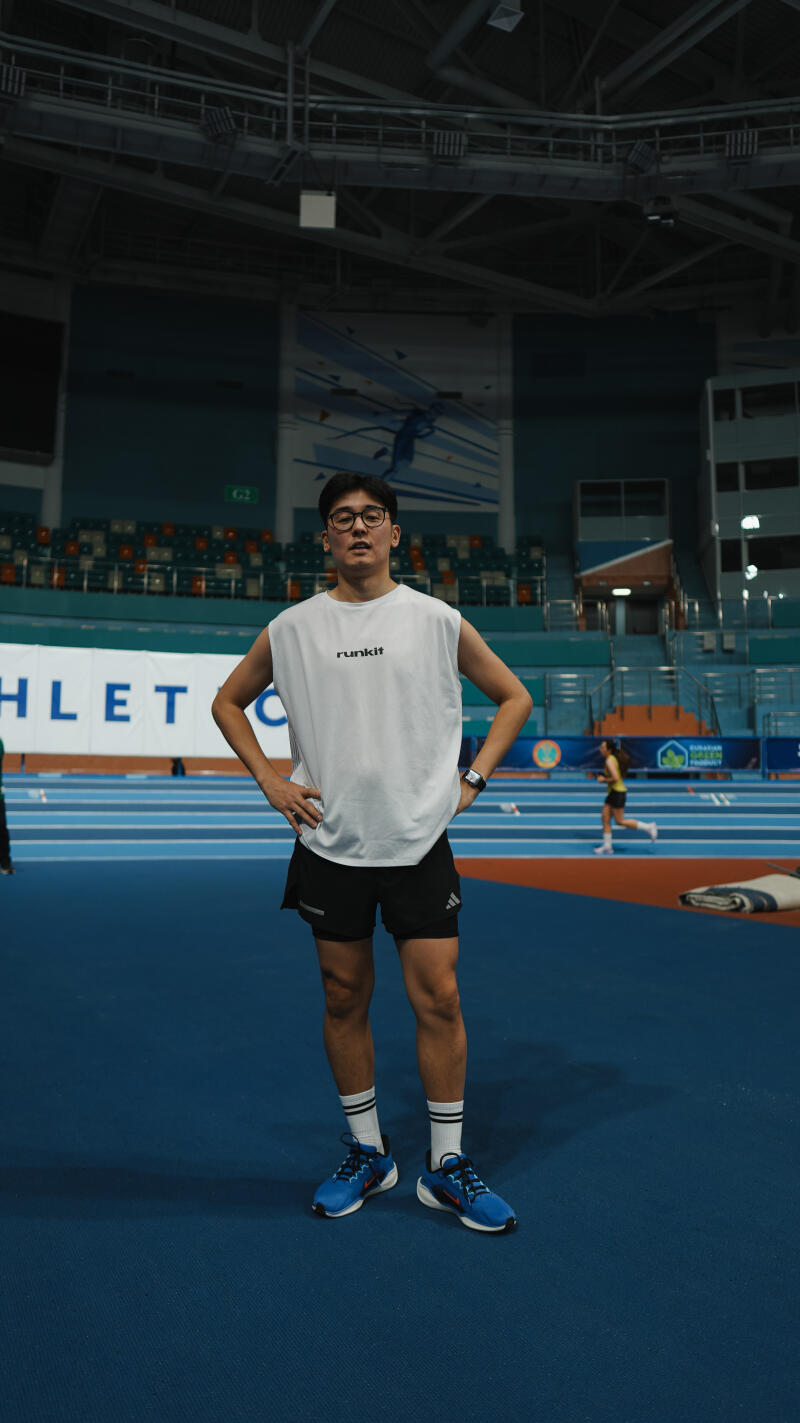 A man with glasses, wearing a white tank top with the word "runkit" on it, black shorts, and blue running shoes, stands with his hands on his hips on a blue running track.