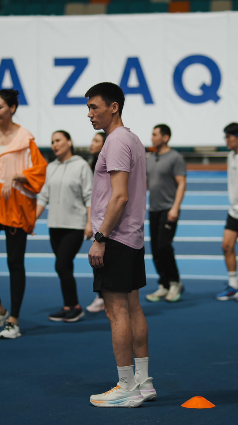 The image shows a man in a pink t-shirt and black shorts standing on a blue track. Behind him are other people, and in the background, there is a sign with the word "AZAQ" in blue letters.