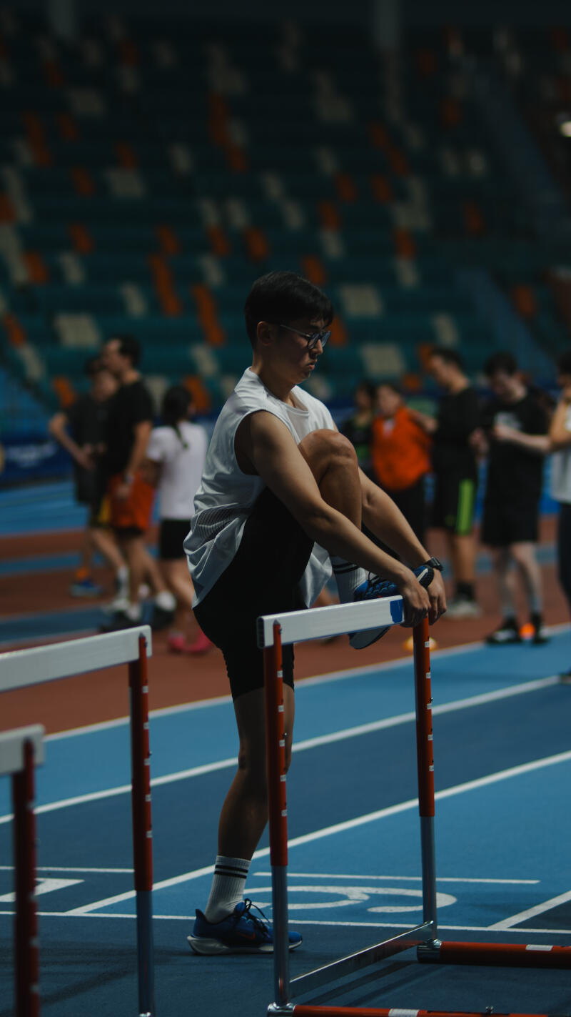 A person in athletic wear stretches their leg on a hurdle on a blue indoor track with other people in the background.