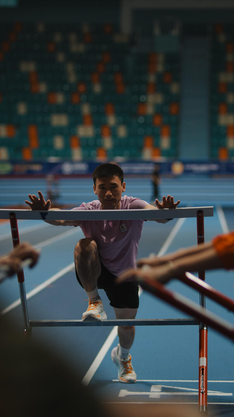 A man in a pink shirt and black shorts is stepping over a hurdle on a blue track, with stadium seating visible in the background.