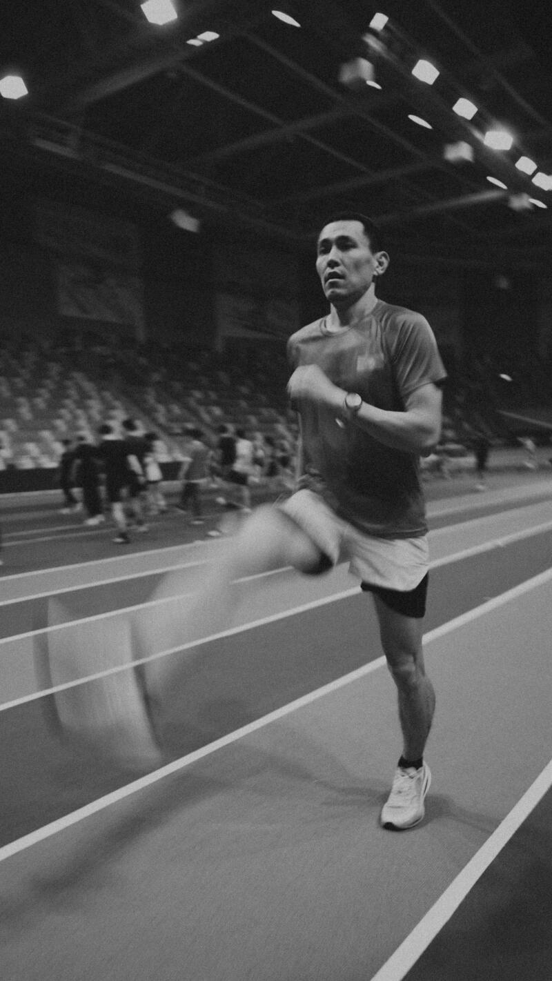 A black and white shot of a man running on an indoor track, wearing a t-shirt, shorts, and running shoes, with spectators blurred in the background.