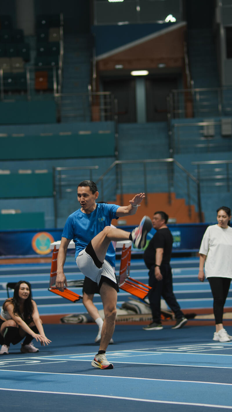 In an indoor athletic setting, a man in a blue shirt and athletic shorts stretches his leg out while standing on a blue track, with other individuals and hurdles visible in the background.