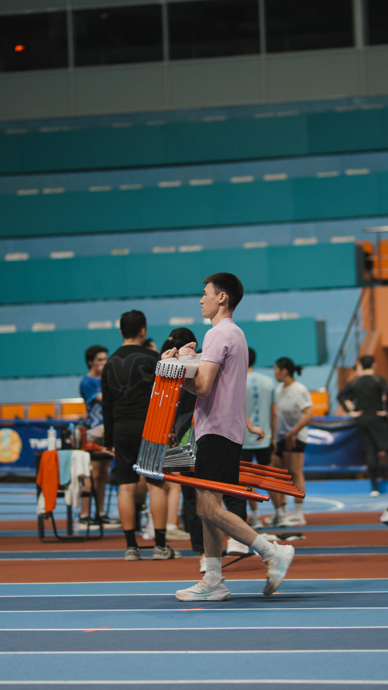 A man in a light purple shirt and black shorts walks across a blue and red track carrying a stack of orange hurdles.
