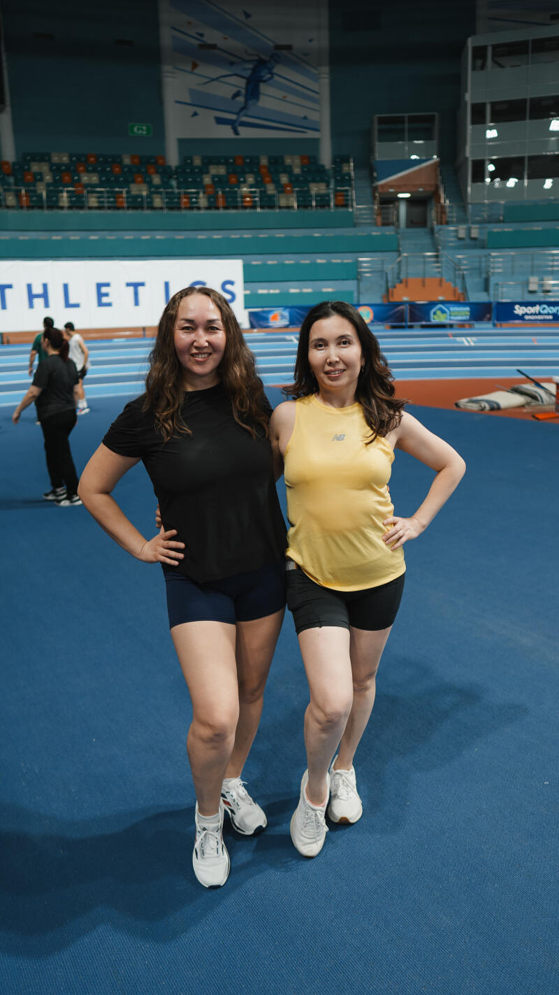 Two women stand side-by-side on a blue indoor track, one wearing a black t-shirt and blue shorts, the other in a yellow tank top and black shorts, both with white sneakers.