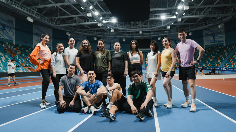 A group of thirteen people are posing on a blue indoor running track with stadium seating visible in the background.