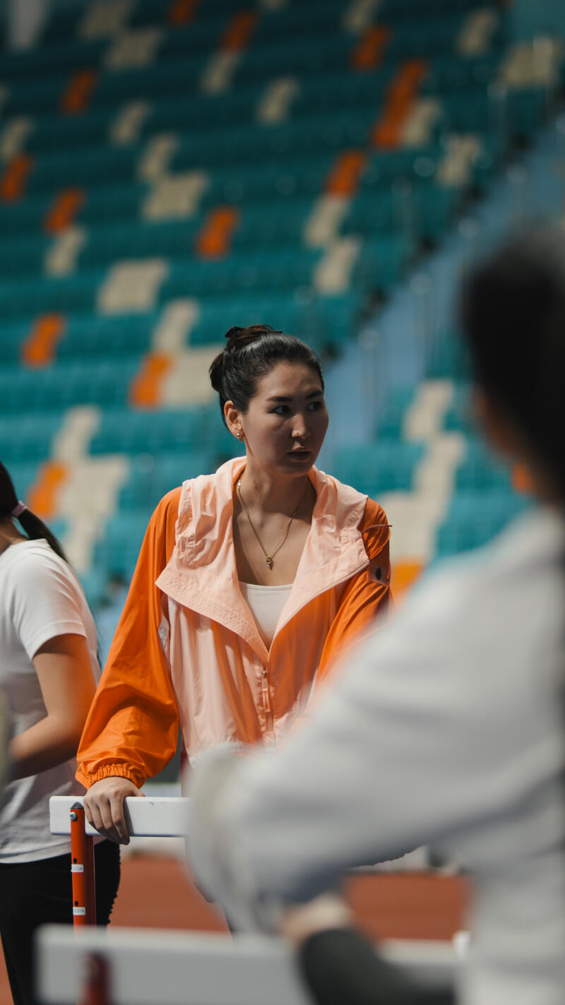 A woman in an orange and pink jacket stands near hurdles in an indoor stadium with blue and white seats.