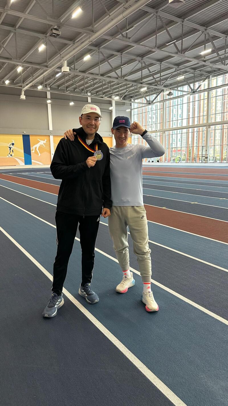 Two men stand on an indoor running track, one wearing a medal and a white baseball cap, the other in a blue baseball cap and athletic wear, posing for a photo.