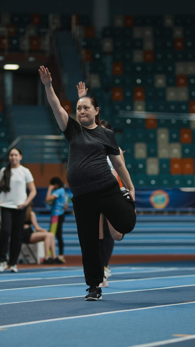 On a blue track, a woman in black pants and a gray shirt stretches her leg while raising her arm, with other people and stadium seating visible in the background.