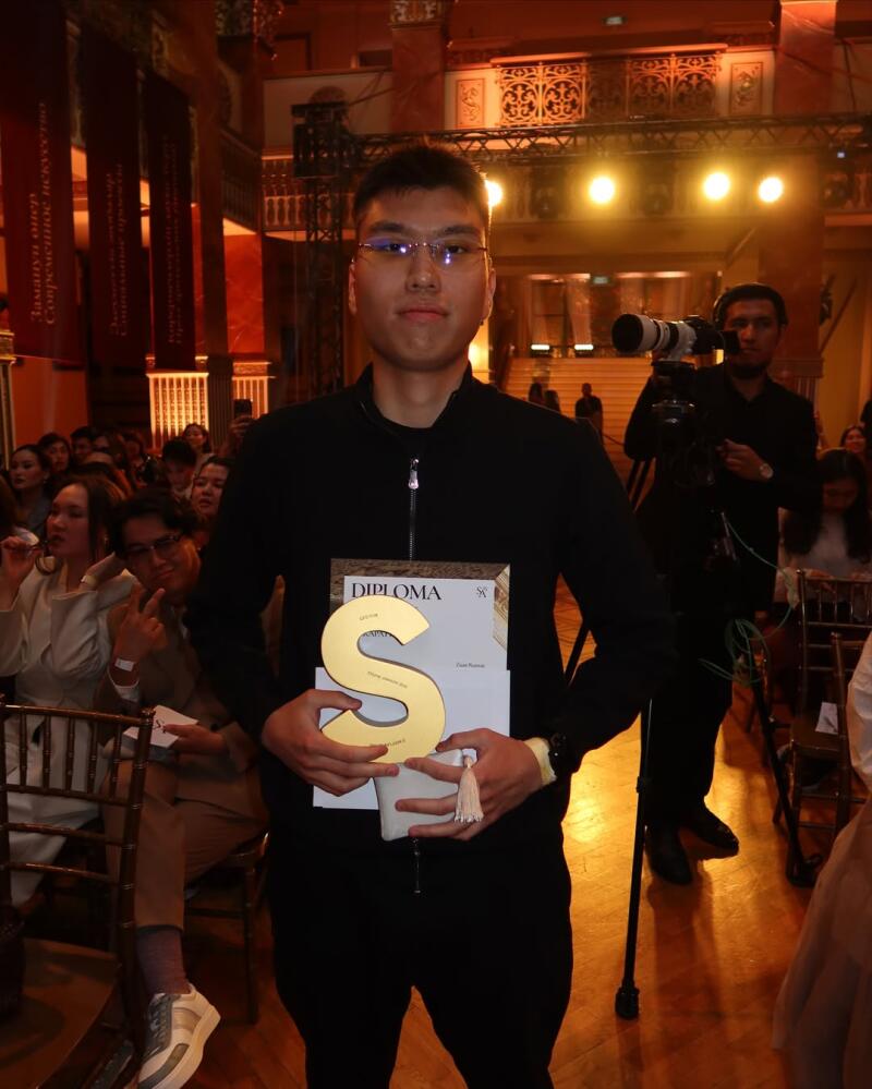 A young man in glasses and a black jacket holds a diploma and a golden S-shaped award, standing in front of an audience and a cameraman.