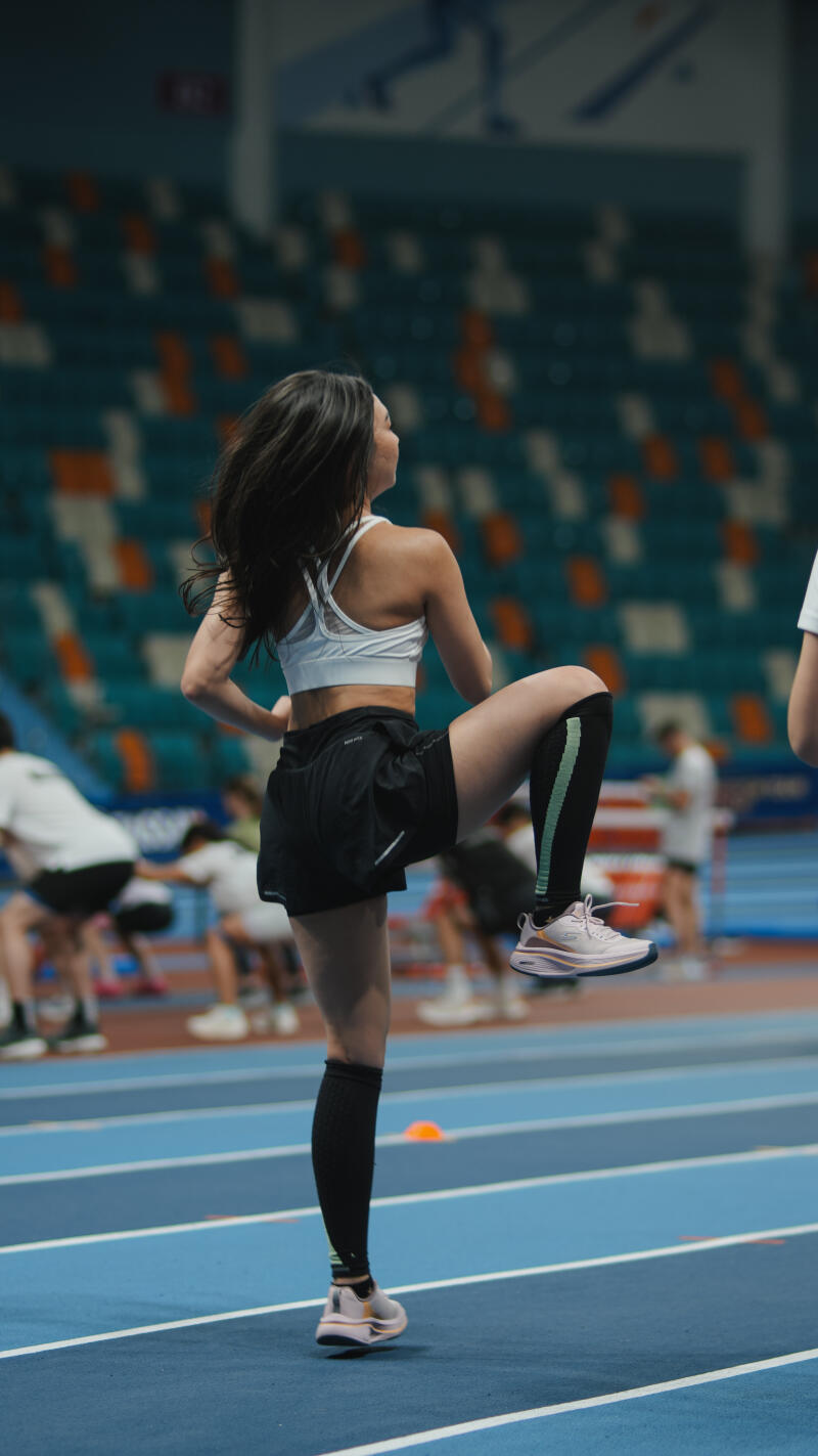A woman in athletic wear is doing a high knee exercise on a blue indoor track, wearing black compression socks, black shorts, a white sports bra, and pink sneakers.