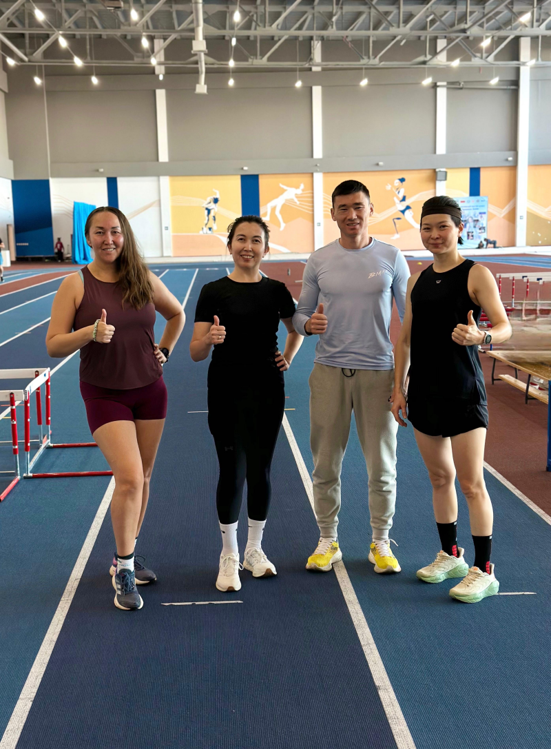 Four people are standing on a blue track with white lines, giving a thumbs-up. They are dressed in athletic wear and sneakers, and there is artwork on the wall behind them.