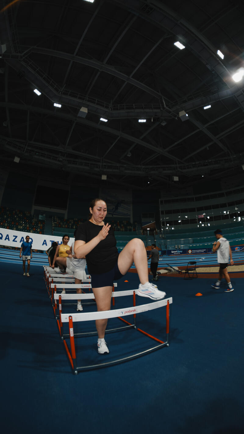 In an indoor stadium, a woman in athletic wear is stepping over a hurdle, with others nearby, and the text "QAZAQ ATLETIKA" visible in the background.