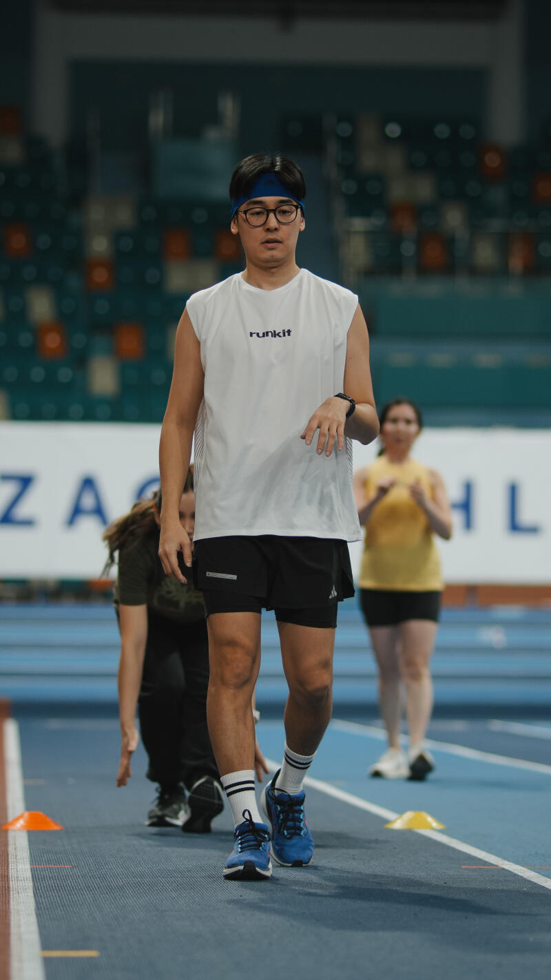 A man with glasses and a blue headband wearing a white tank top with the word "runkit" on it, black shorts, white socks, and blue sneakers is walking on a blue track with other people in the background.