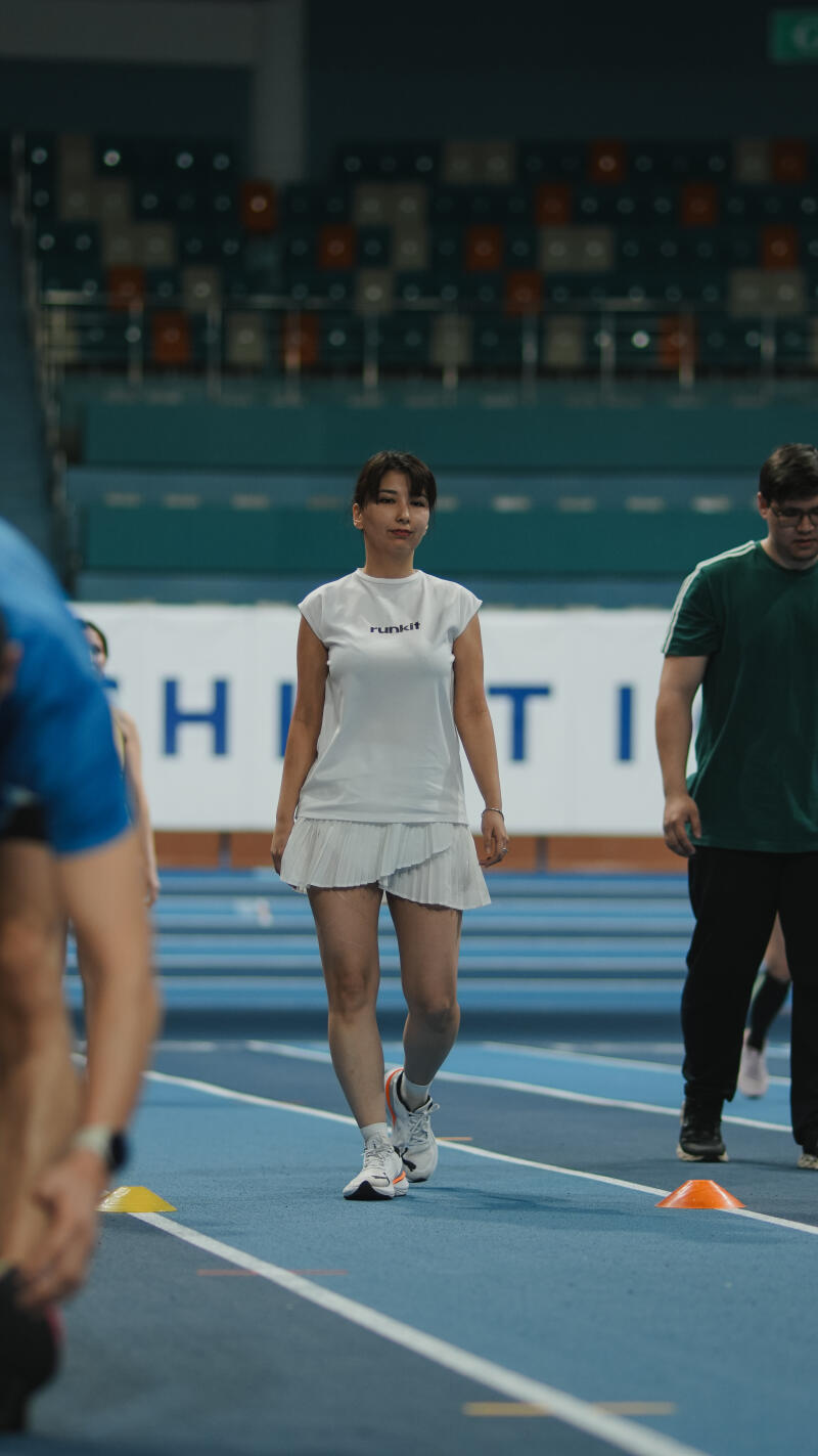 A woman in a white "runkit" t-shirt and skirt is walking on a blue track in an indoor stadium.