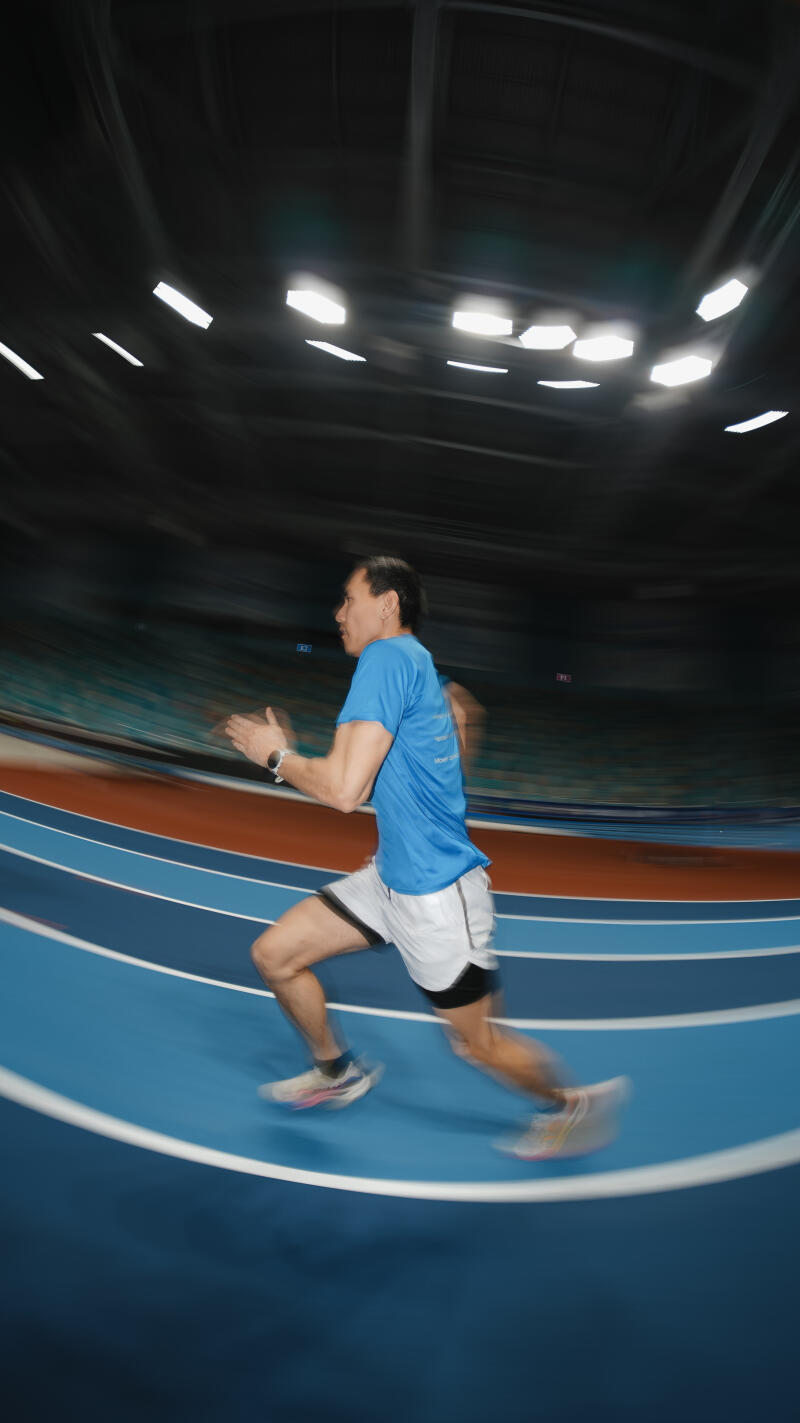 A man in a blue shirt and white shorts is running on a blue track in an indoor stadium with blurred motion.