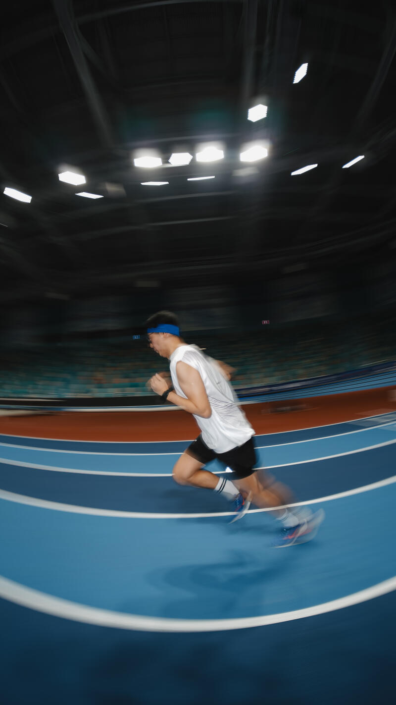A man wearing a blue headband, white shirt, and black shorts is running on a blue track with white lines in an indoor stadium.