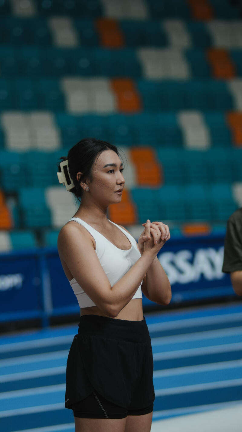 A woman with dark hair pulled back with a white clip is wearing a white top and black shorts, standing in front of stadium seating with blue, white, and orange colors.