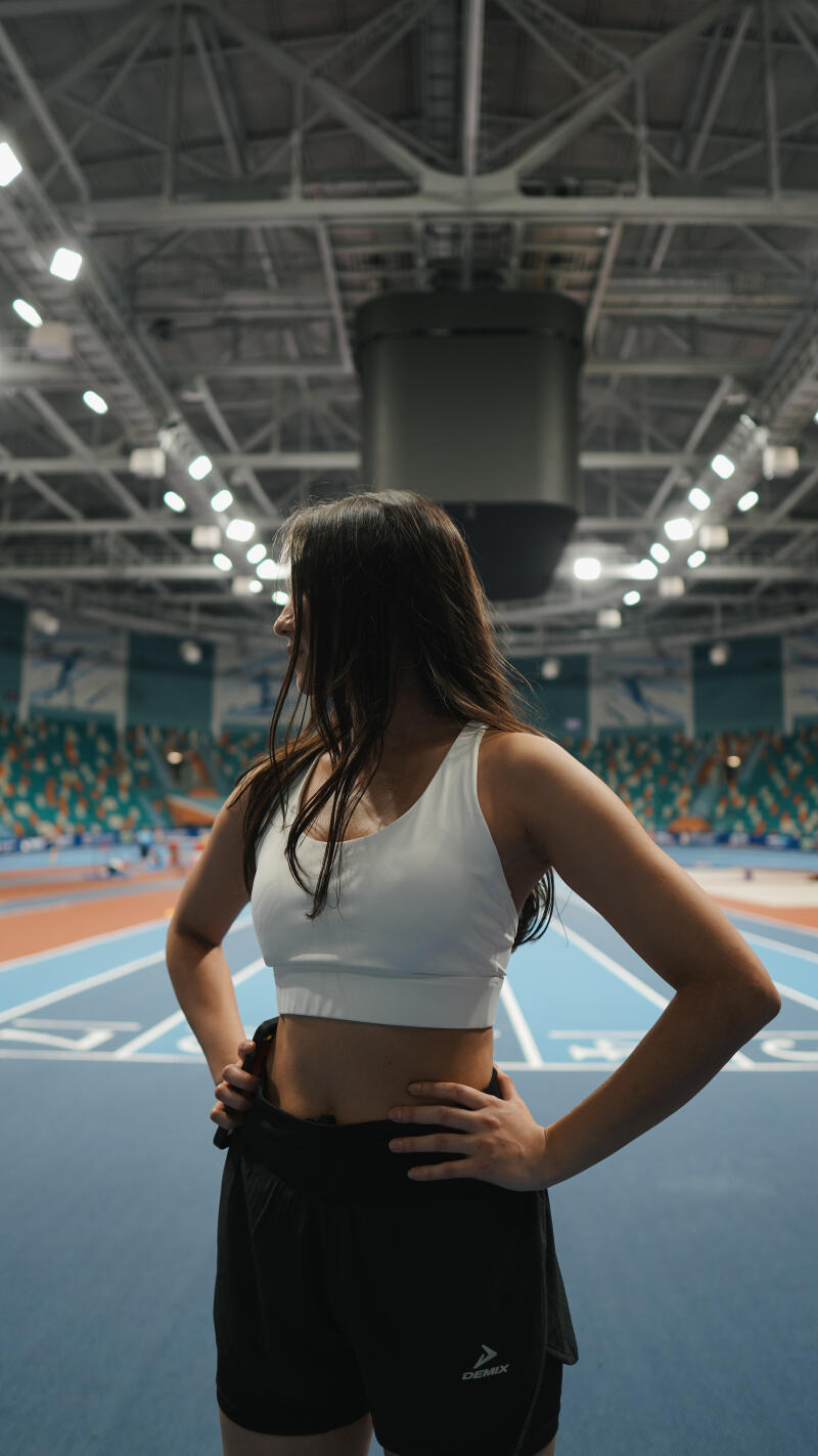 A woman in a white sports bra and black shorts with 'DEMIX' visible on the shorts stands in an indoor stadium with running tracks and spectator seating.