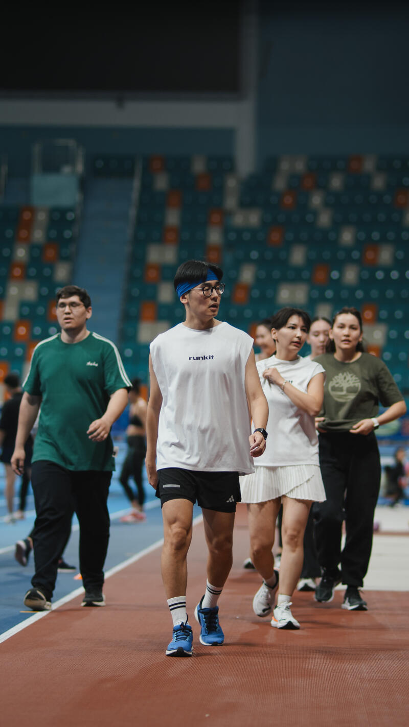 Several people are walking on a track, including a man in a white 'runkit' tank top and blue headband.