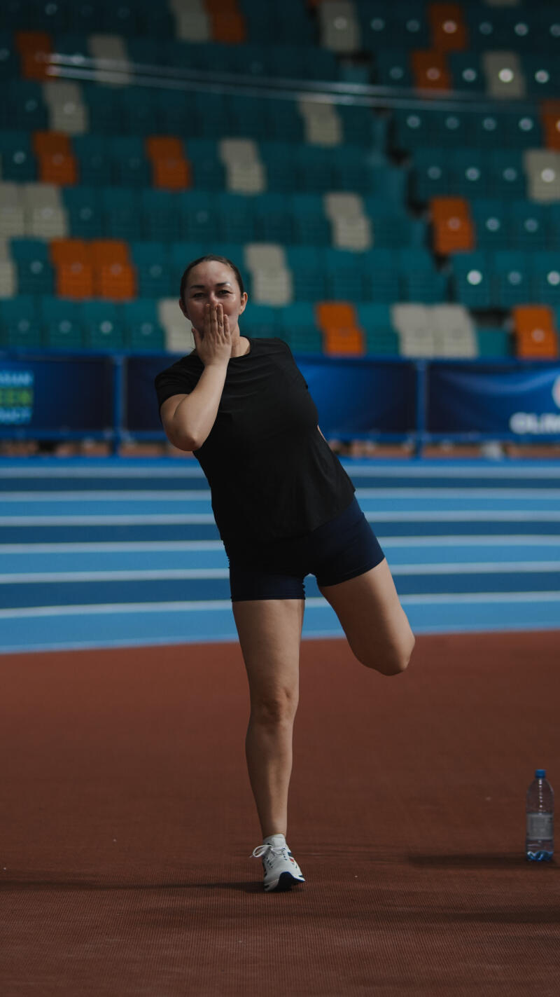 A woman in a black t-shirt and blue shorts is stretching her leg on a running track in front of stadium seating.