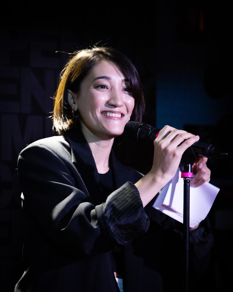 A woman in a black blazer smiles while holding a microphone and a piece of paper.
