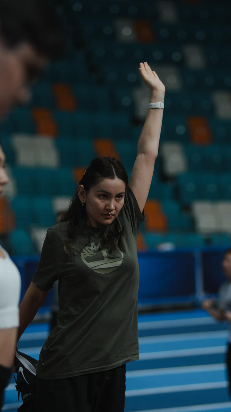 A woman in an olive green T-shirt stretches her arm upward while holding her leg in her hand.