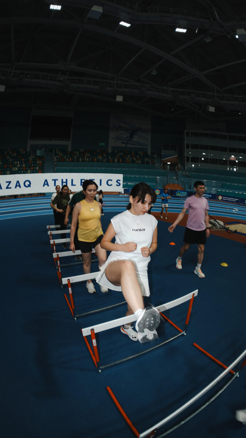 A group of people, including a woman in a white "runkit" t-shirt and skirt, are stepping over small hurdles on a blue track inside a large indoor stadium with the text "AZAQ ATHLETICS" visible in the background.