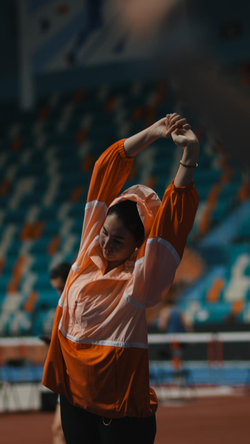 A woman in an orange and pink jacket stretches her arms above her head in front of stadium seating.