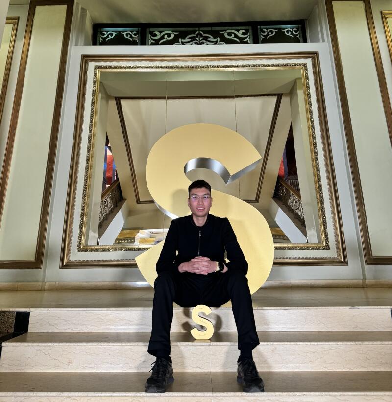 A man in a black suit sits on stairs with a large gold "S" behind him and a smaller gold "S" in front.