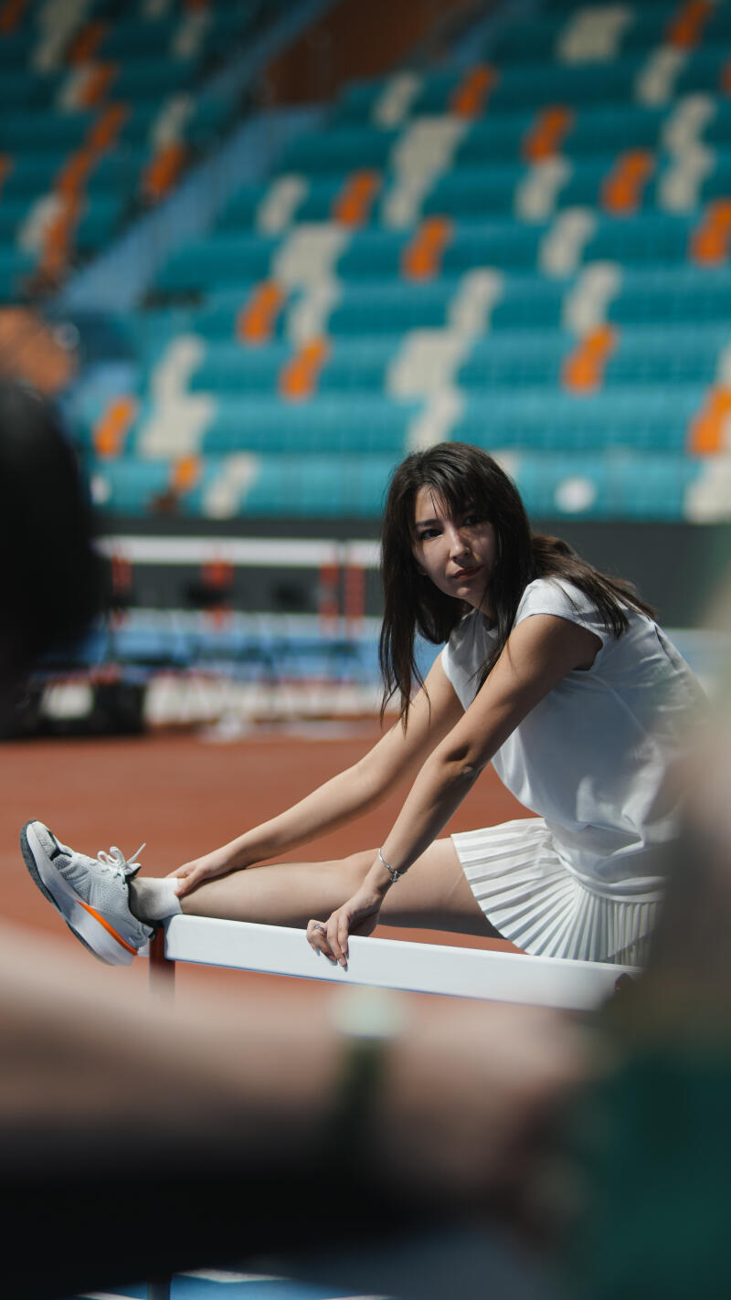 A woman in a white shirt and skirt stretches her leg on a white railing in front of stadium seating.