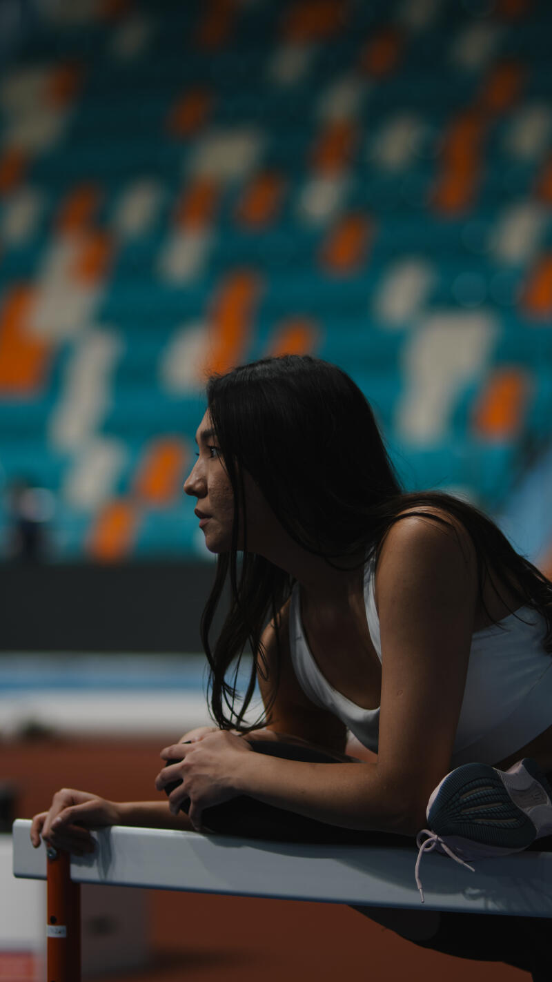 A woman with long dark hair, wearing a white tank top, is leaning on a hurdle, with a running shoe beside her, in front of blurred stadium seating.