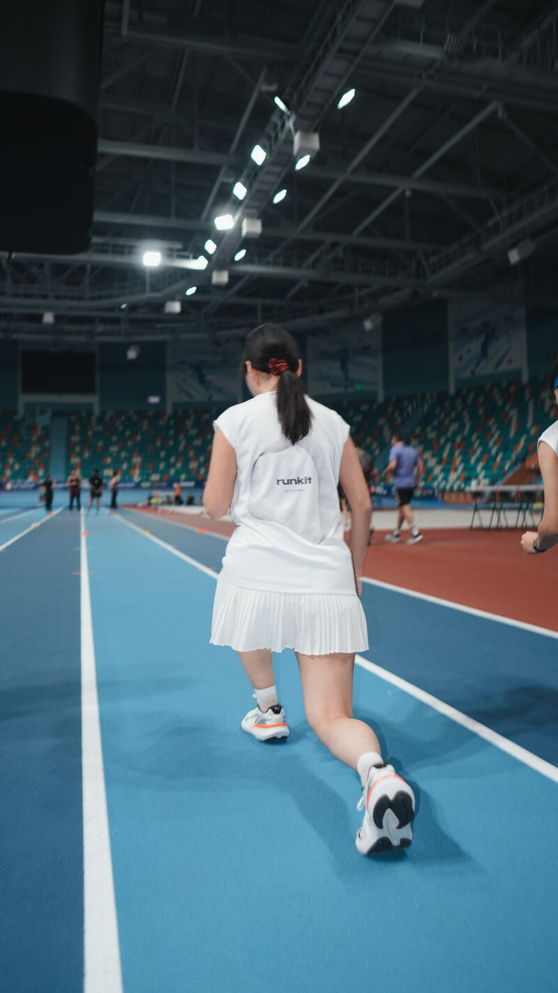 A person in a white shirt and pleated skirt is walking on a blue track, with the word "runkit" visible on the shirt.