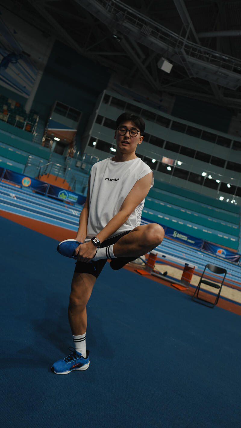 A man wearing a white tank top with "runk!" printed on it, black shorts, and blue running shoes is stretching his leg on a blue track.