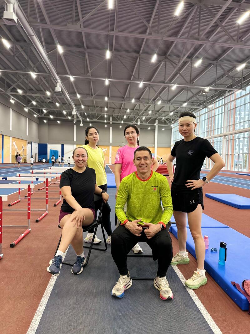 Five people pose indoors in athletic wear, with hurdles and a running track in the background.