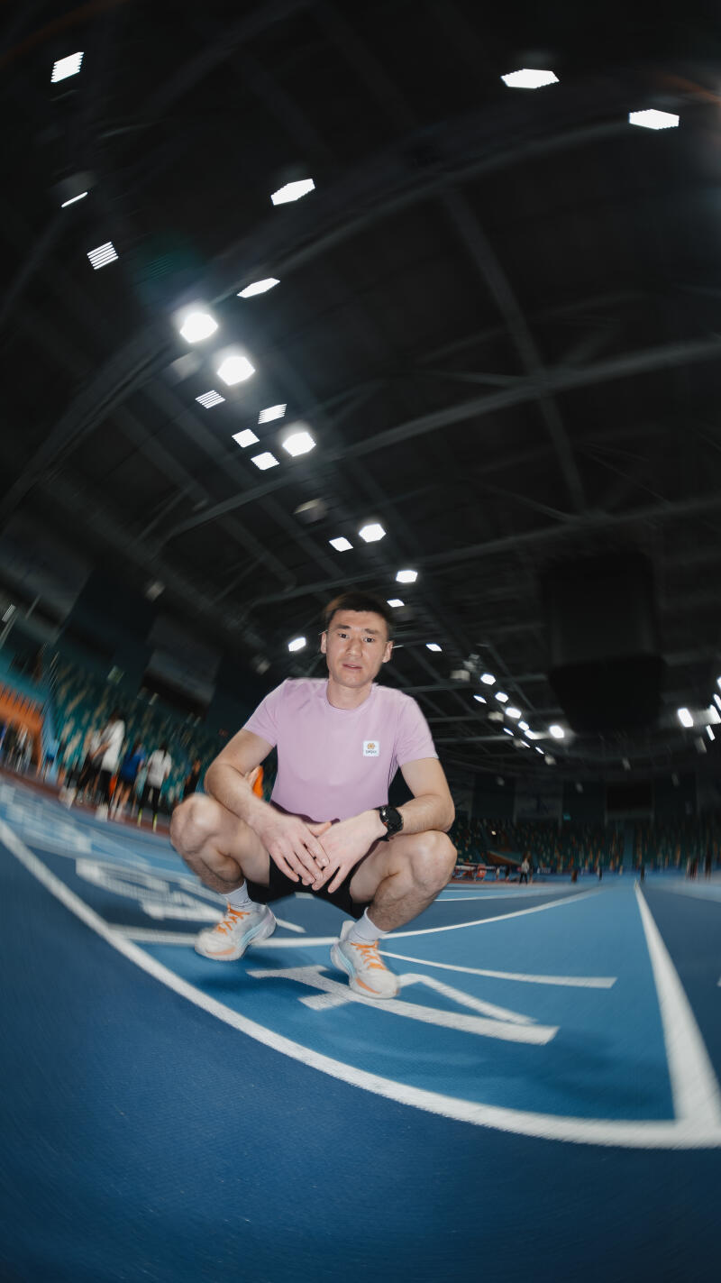 A man in a pink t-shirt and black shorts is squatting on a blue running track, wearing orange and white sneakers and a watch on his left wrist.