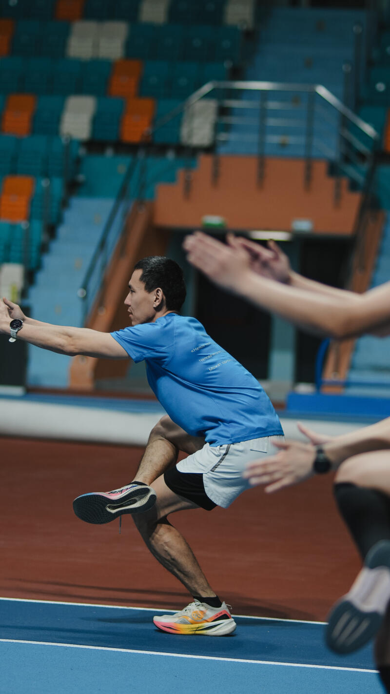 A man in a blue shirt, shorts, and running shoes is exercising, with one leg extended in front and the other bent underneath him, on a blue and red track in front of stadium seating, as others around him clap their hands.