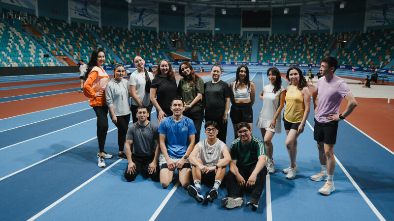 A group of thirteen people are posing for a photo on an indoor running track with stadium seating in the background.