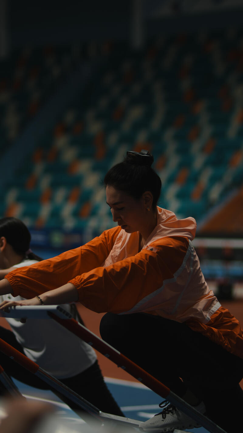 A woman in an orange jacket is stretching at a track and field event with stadium seating in the background.